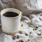 Small white mug of Cardamom Coffee, surrounded with cardamom pods