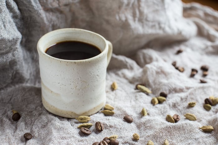 Small white mug of Cardamom Coffee, surrounded with cardamom pods