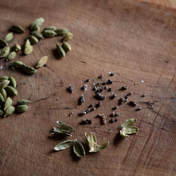 wooden background with green cardamom pods, some of which have been split open to expose their little black seeds