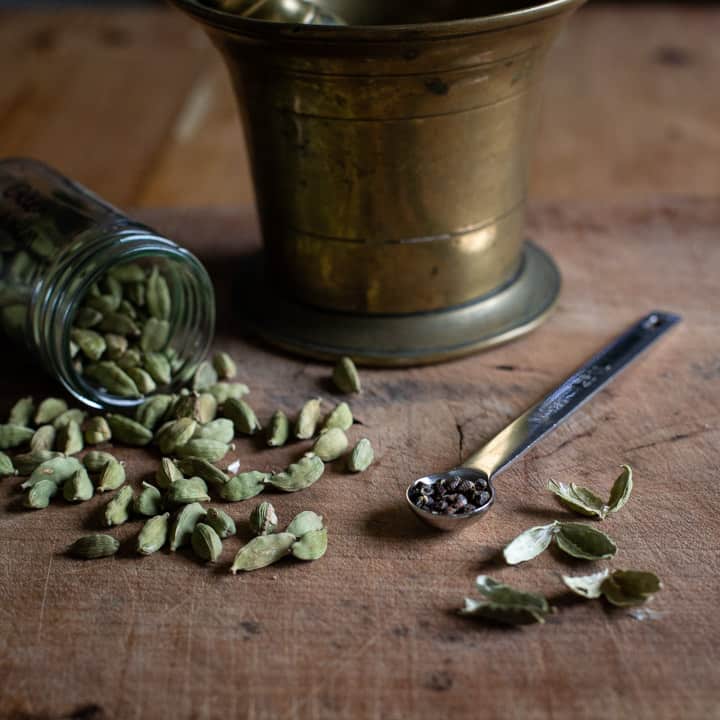 wooden background with cardamom pods and a teaspoon full of little black cardamom seeds