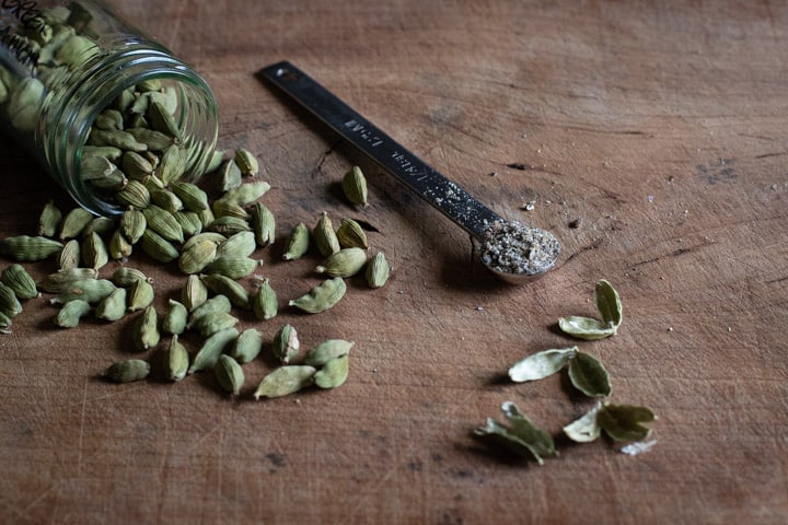 wooden background with cardamom pods and a teaspoon full of freshly ground cardamom seeds