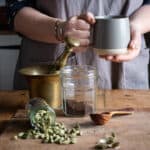 womands hands holding a grey mug of hot cardamom coffee over a messy scene of cardamom pods and coffee making paraphenalia