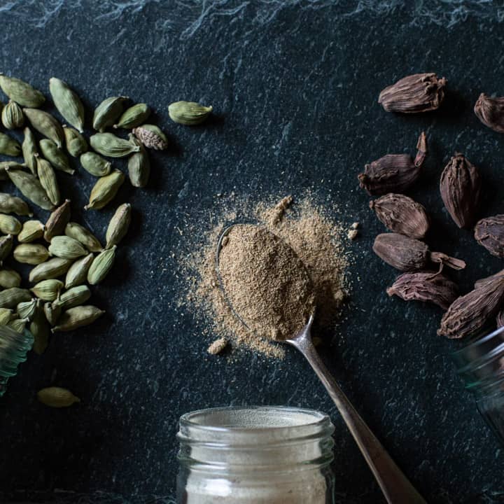 black slate background with whole green cardamom pods, whole black cardamom pods and a silver teaspoon with ground cardamom spice