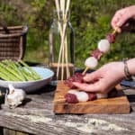 womans hands threading steak and scallops into wooden skewers in a rustic garden setting