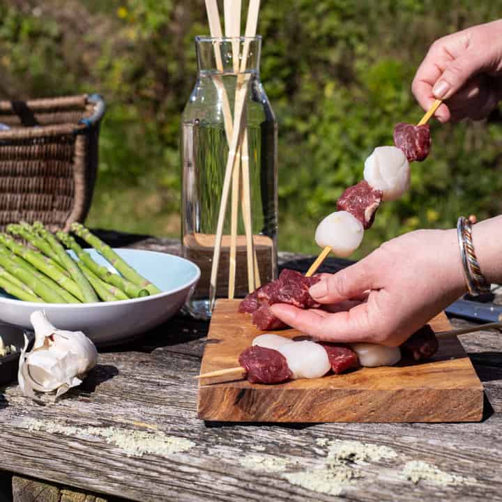 womans hands threading steak and scallops into wooden skewers in a rustic garden setting