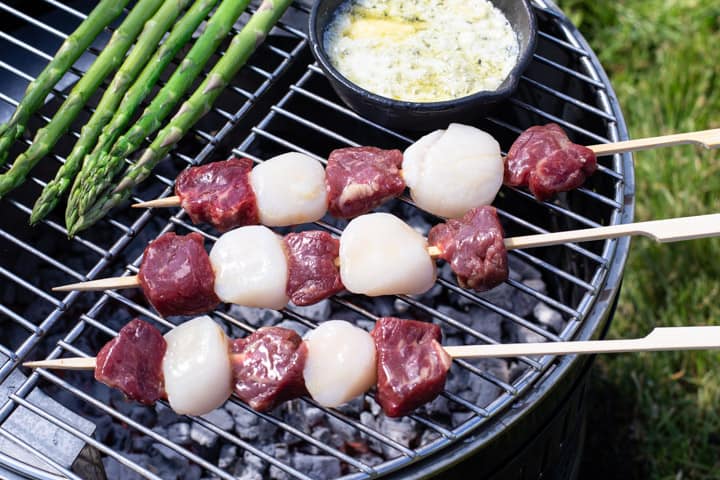 3 sirloins steak and scallop kebabs cooking on a garden BBQ with asparagus spears and garlic butter cooking in small cast iron pan