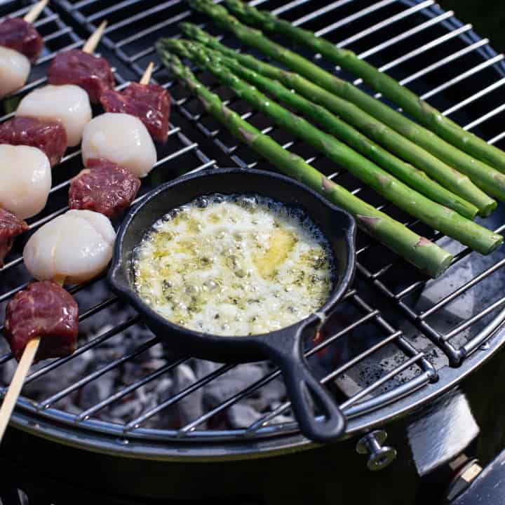 small black cast iron skillet filled with bubbling garlic butter on top of a BBQ with surf and turf kebabs and fresh asparagus spears