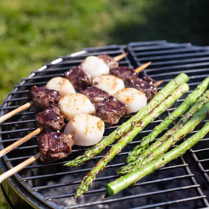 3 surf n turf kebabs cooking with asparagus on a silver and black grill with green grass in the background