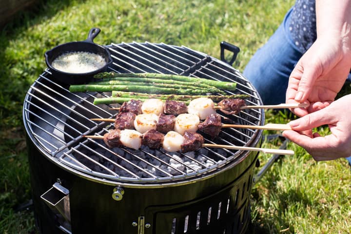 womans hands turning over surf and turf skewers on a BBQ grill with asparagus and a pot of garlic butter