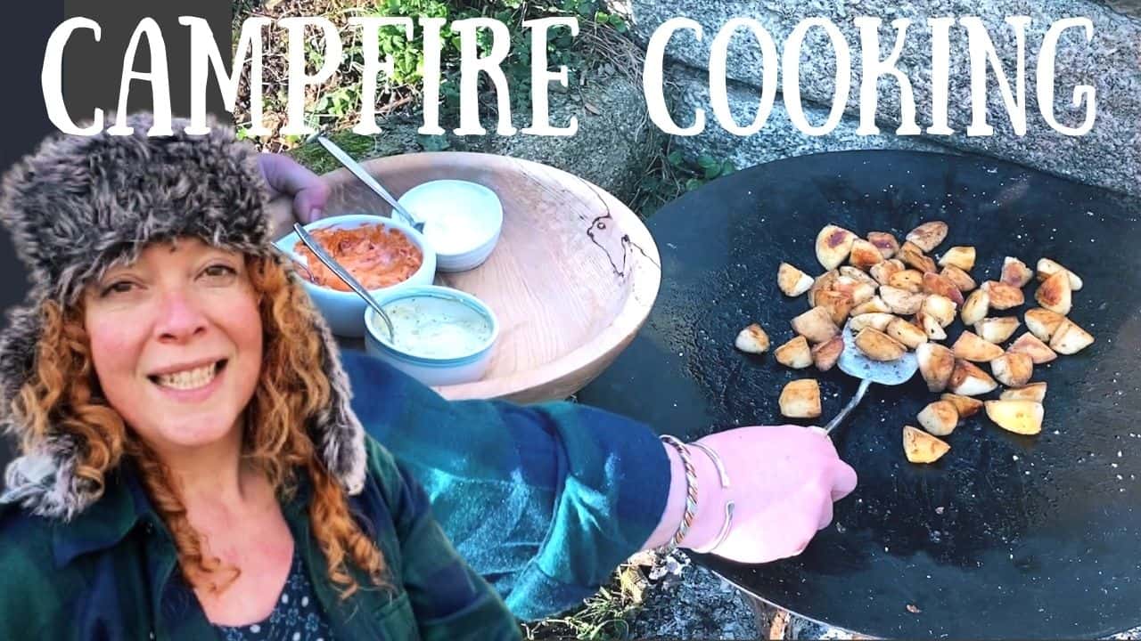 woman with red hair and black hat in front of a campfire where saute potatoes are cooking in a large black metal skillet