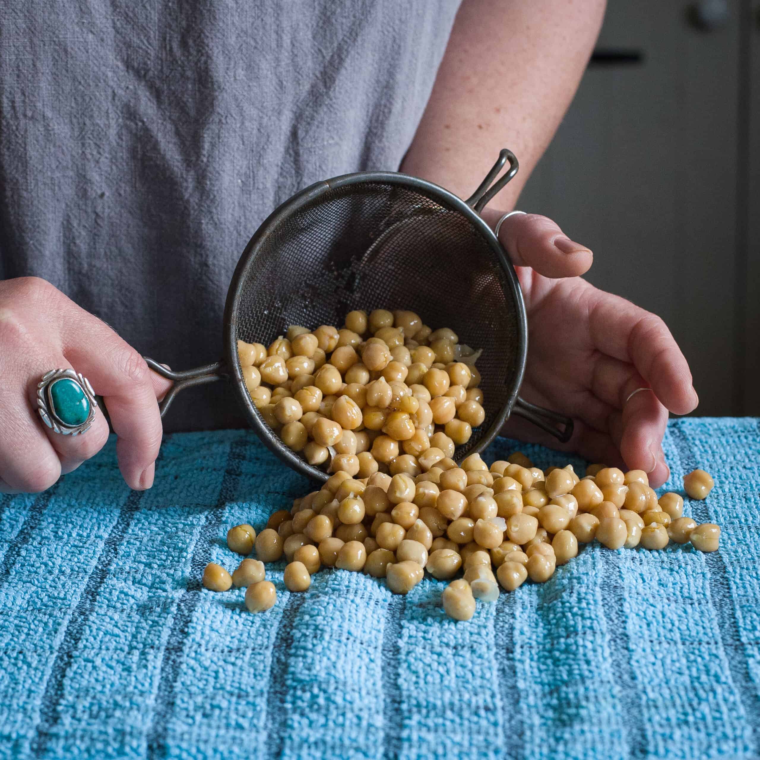 Woman’s hands tipping drained chickpeas from an old fashioned sieve onto a blue tea towel to dry