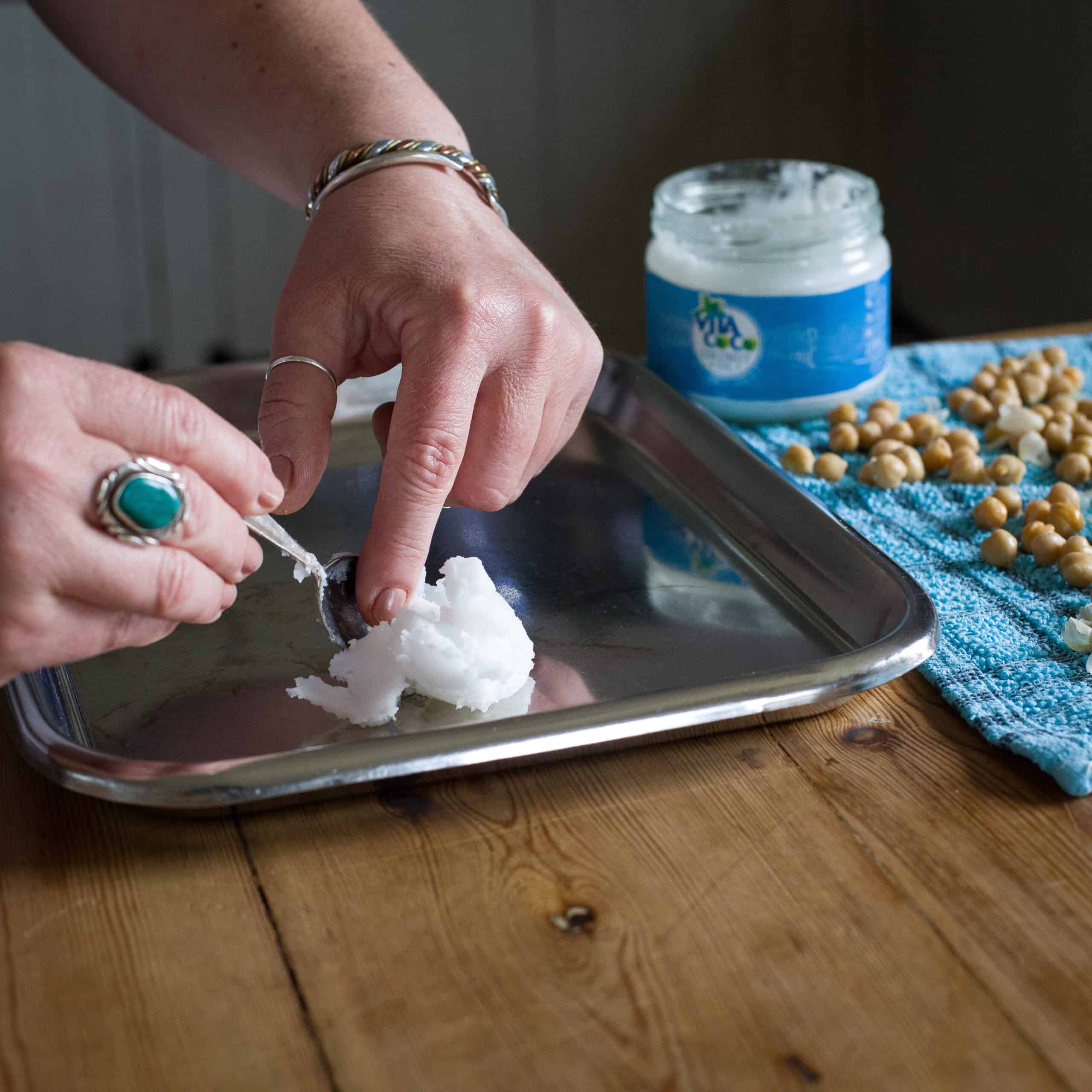 Woman’s hands spooning a tablespoon of coconut oil onto a stainless steel baking sheet to roast chickpeas