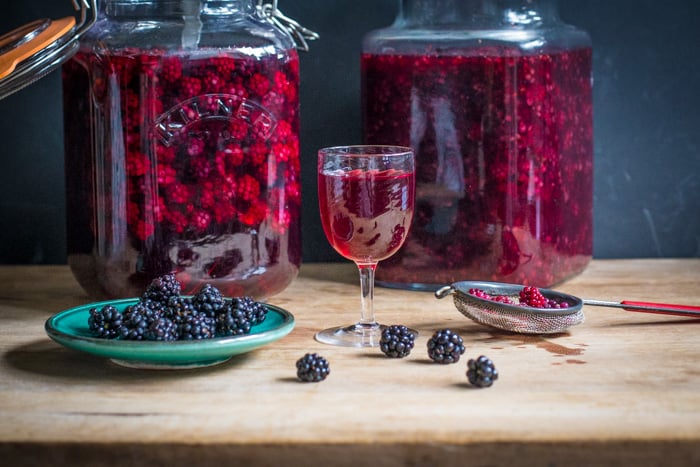 Blackberry Flavoured Vodka in a small wine glass and 2 kilner jars with a dish and a sieve both containing blackberrys