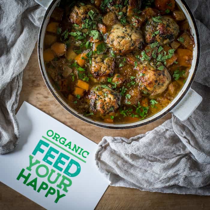 Ox Cheek Stew with Nettle Dumplings in a casserole dish on a wooden tavle with a cloth and a sign reading Organic Feed Your Happy