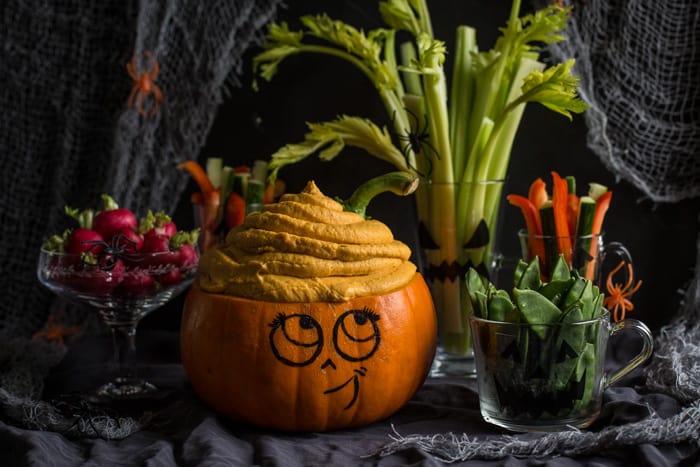 Cute Halloween pumpkin head overflowing with pumpkin dip, surrounded by vegetable crudites