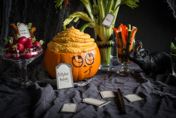 Healthy Halloween recipes includes dip labelled pumpkin brains, served in a scooped out pumpkin with a smiley face drawn onto it and crudites in glasses around it