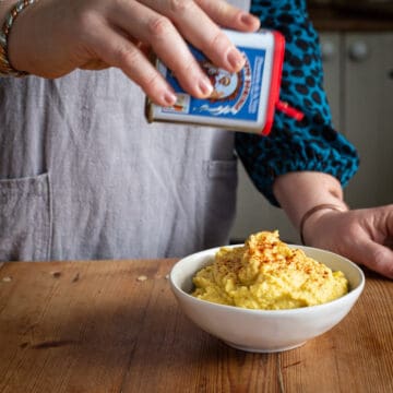 Woman’s hands sprinkling smoked paprika from a white tub with a red lid onto a bowl of homemade pumpkin dip
