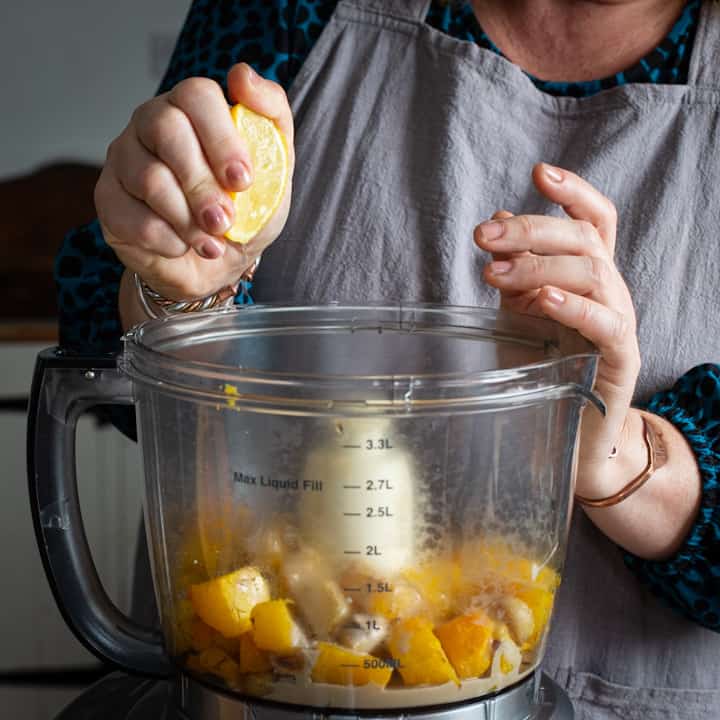 Woman in grey squeezing half a lemon into a food processor filled with pieces of roast pumpkin flesh