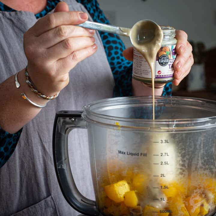 Woman in grey pouring a spoon of tahini into a food processor filled with cubes of pumpkin flesh