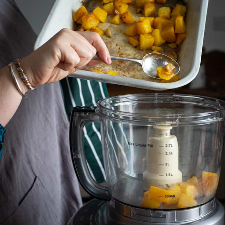 Woman in grey scraping chunks of roast pumpkin into a food processor