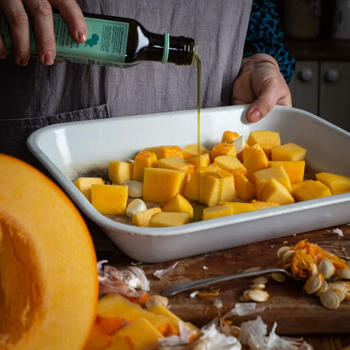 Woman in grey pouring avocado oil into a baking tray of chopped pumpkin