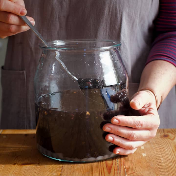 Woman in grey holding a spoon of sloes over a large glass jar of brewing sloe vodka liqueur