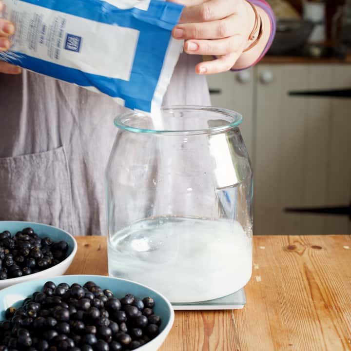 Woman in grey pouring sugar from a blue and white bag into a large glass jar of vodka on a wooden counter top