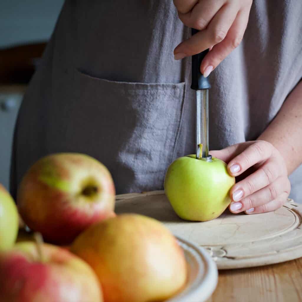 Woman using a silver apple corer to remove the cores from several apples on a wooden chopping board