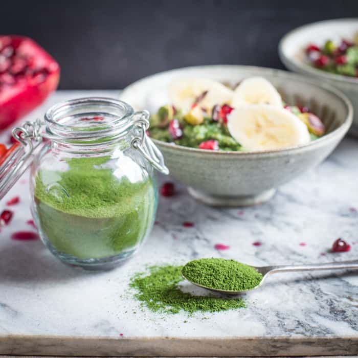 Small glass jar of dehydrated DIY Green Vegetable Powder with a bowl of porridge on a marble slab