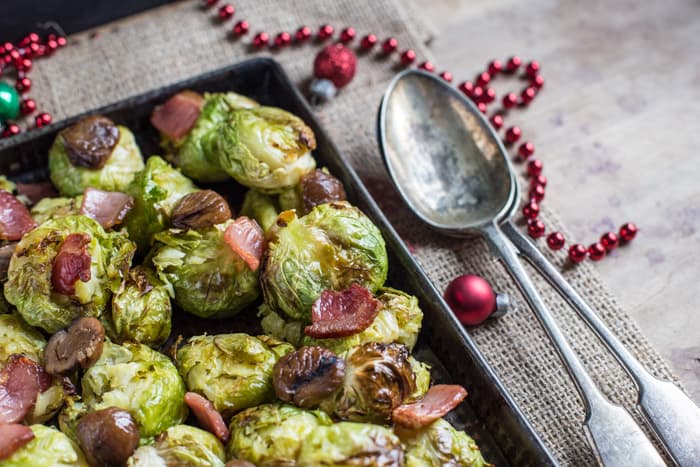 Smashed Sprouts with Bacon and Chestnuts in a roasting tin with a seving spoon beside them and Christmas decorations on the table