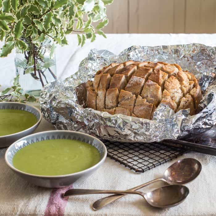 two bowls of pea and mint soup with spoons, soup container, foliage and loaf of garlic hedgehog sourdough bread