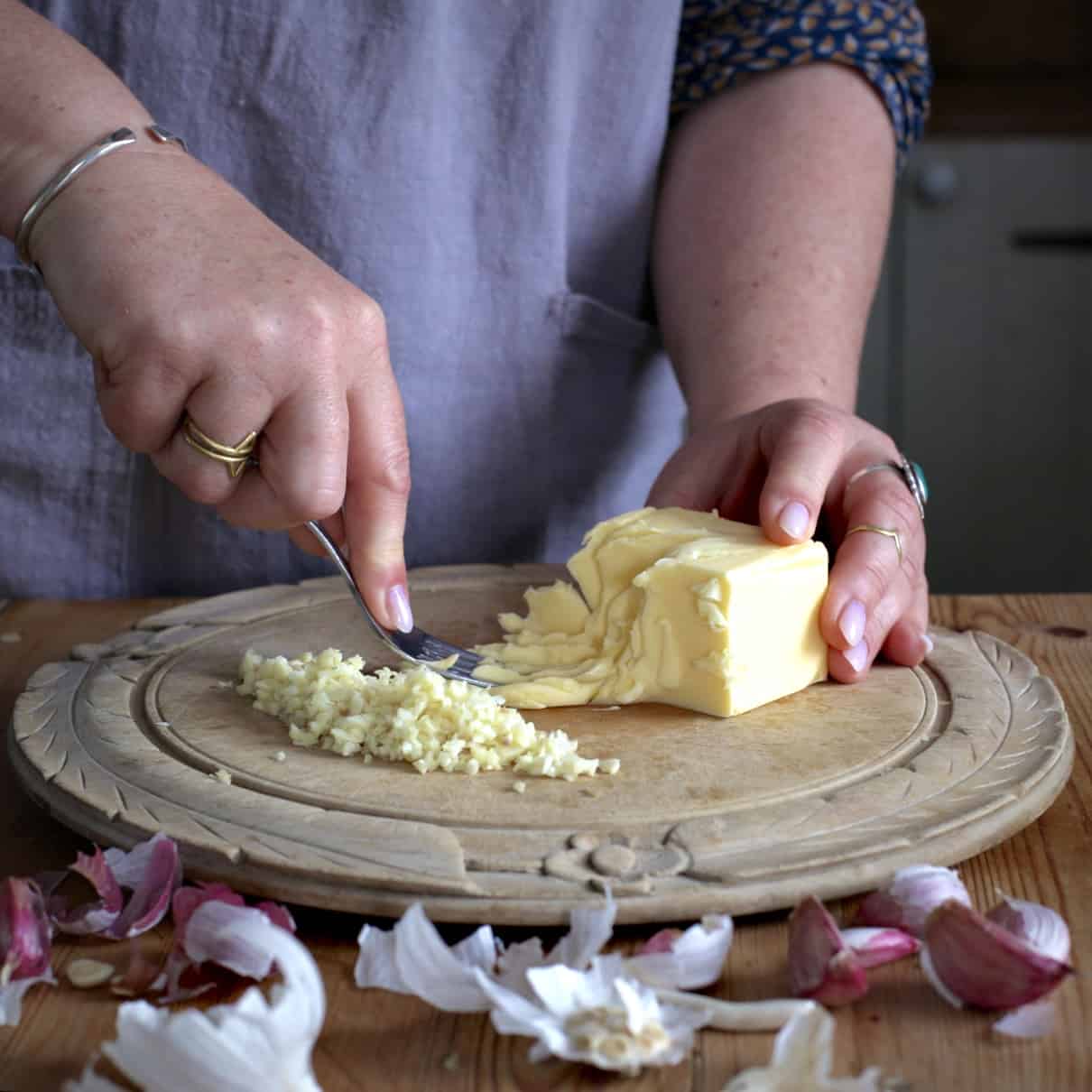 Womans hands mashing butter on a wooden board next to a pile of minced garlic