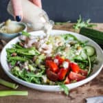 Small glass bottle of tahini sauce surrounded by a vibrant mix of salad vegetables, sauerkraut and olives