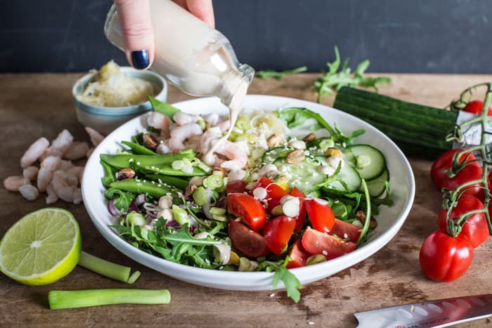 Small glass bottle of tahini sauce surrounded by a vibrant mix of salad vegetables, sauerkraut and olives