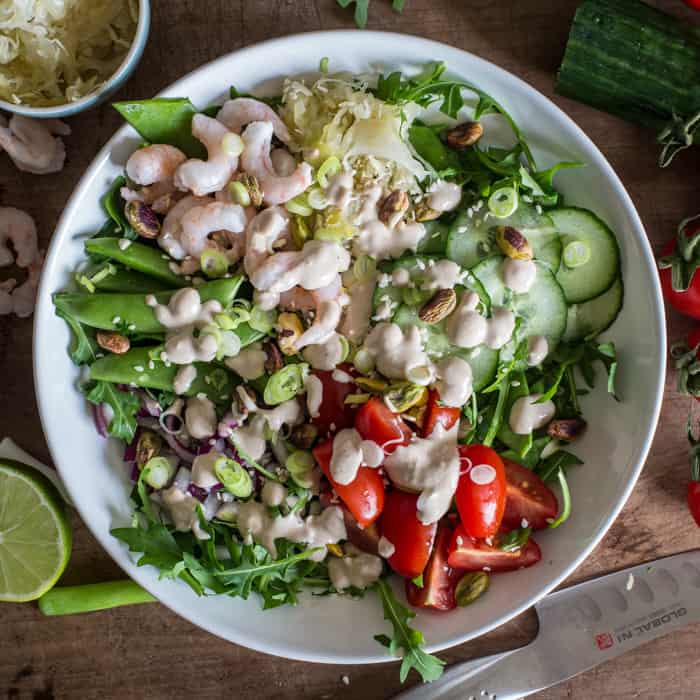 Small glass bottle of tahini sauce surrounded by a vibrant mix of salad vegetables, sauerkraut and olives