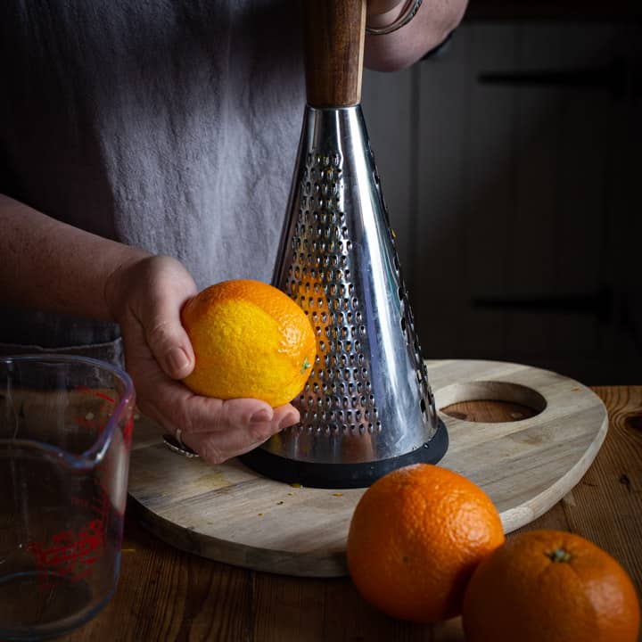 woman zesting an orange on a large metal grater over a wooden chopping board