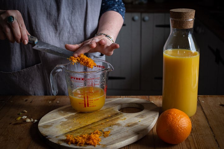 womans hands scraping orange zest from the blade of a knife into a glass jug of orange juice