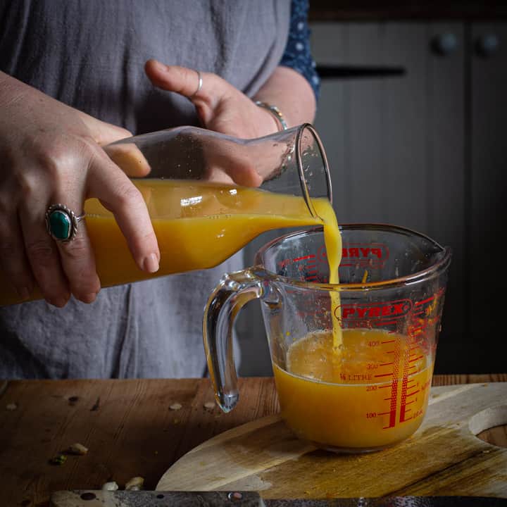 woman in grey pouring orange juice into a glass measuring jug from a glass carafe