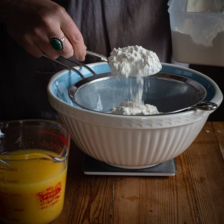 womans hand spooning flour into a blue and white mixing bowl on a wooden kitchen counter