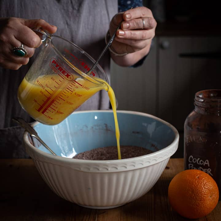 woman in grey pouring orange juice into a blue and white bowl of muffin batter