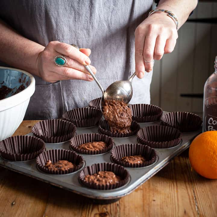 womans hands spooning chocolate muffin batter into brown muffin cases in a silver baking tray