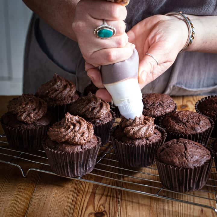 womans hand piping chocolate buttercream onto chocolate muffins