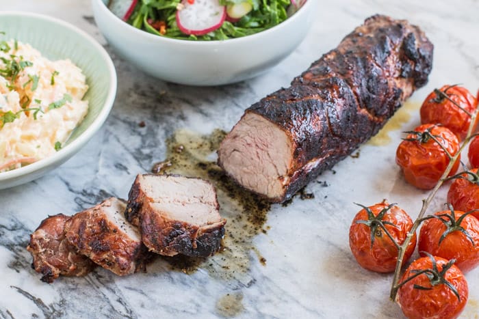 Juicy bbq pork tenderloin sliced on marble slab with bowls of salad and roast tomatoes