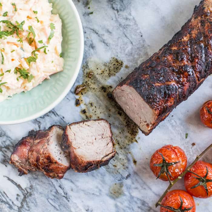 Juicy bbq pork tenderloin sliced on marble slab with bowls of salad and roast tomatoes