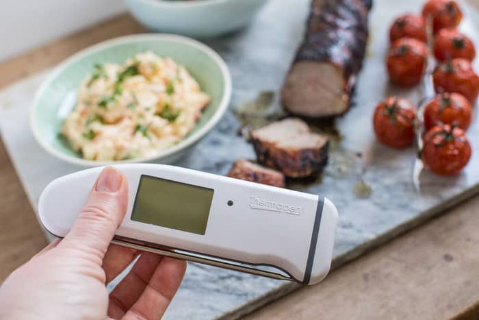 Juicy bbq pork tenderloin sliced on marble slab with bowls of salad and roast tomatoes and a digital meat thermometer