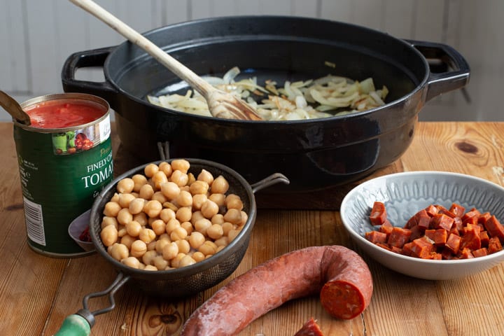 black pan with friend onions a sieve of drained chickpeas 1/2 a chorizo sausages and a can of chopped tomatoes sitting on a wooden kitchen bench