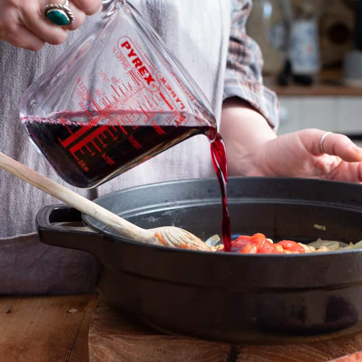 womans hands pouring red wine into a black cast iron casserole dish of chicken chorizo stew