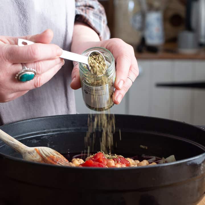 womans hands spooning dried oregano into a cast iron pan of chicken stew