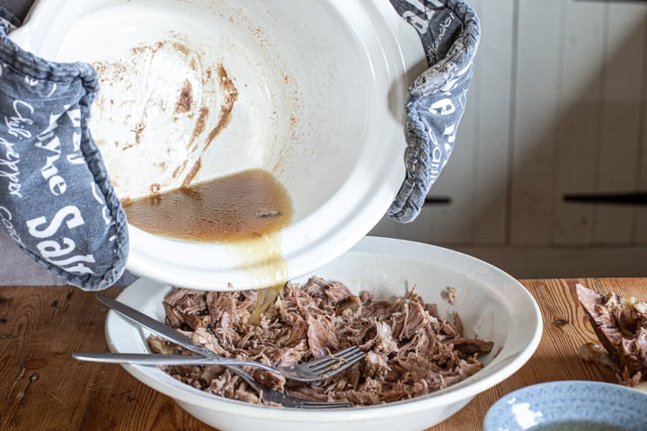 white slow cooker bowl of meat juices being poured over a white bowl of shredded pulled lamb