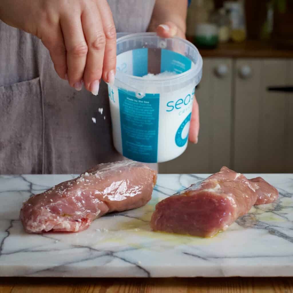 Womans hands sprinkling Cornish sea salt from a white and blue tub onto a pork tenderloins on a marble board